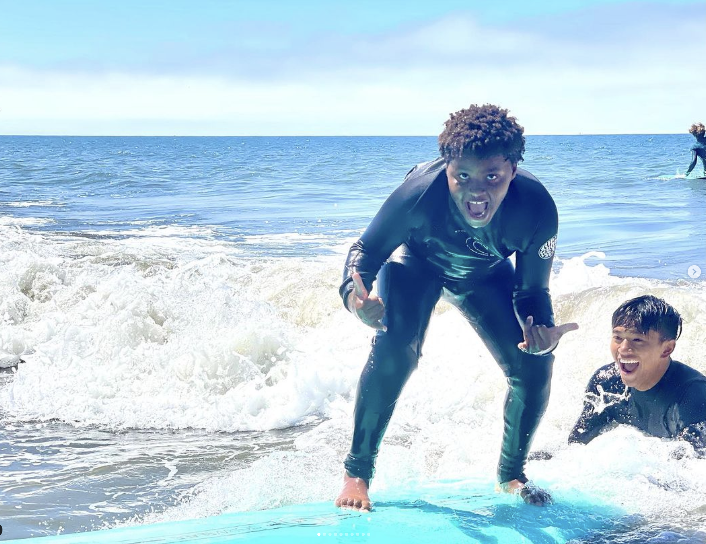 A young participant grinning as he surfs a wave while an instructor watches