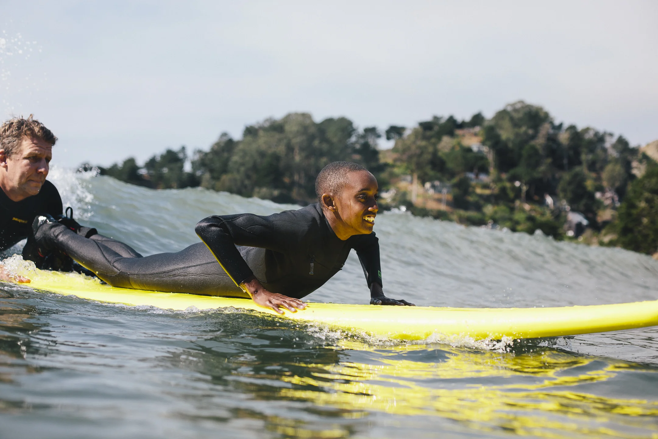 A young participant paddling a yellow surfboard into a wave with an instructor alongside
