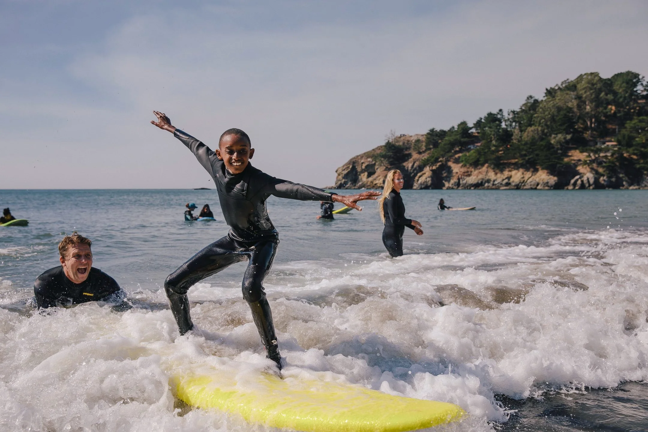 A young boy riding a yellow surfboard with arms spread as an instructor cheers in the water