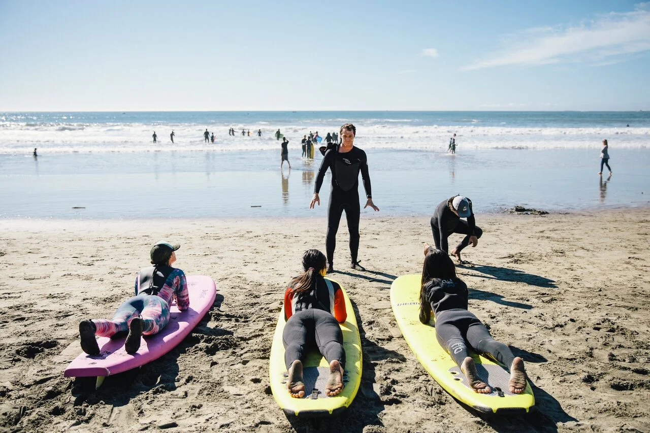 A male instructor demonstrating pop-ups to three girls lying on surfboards on the sand
