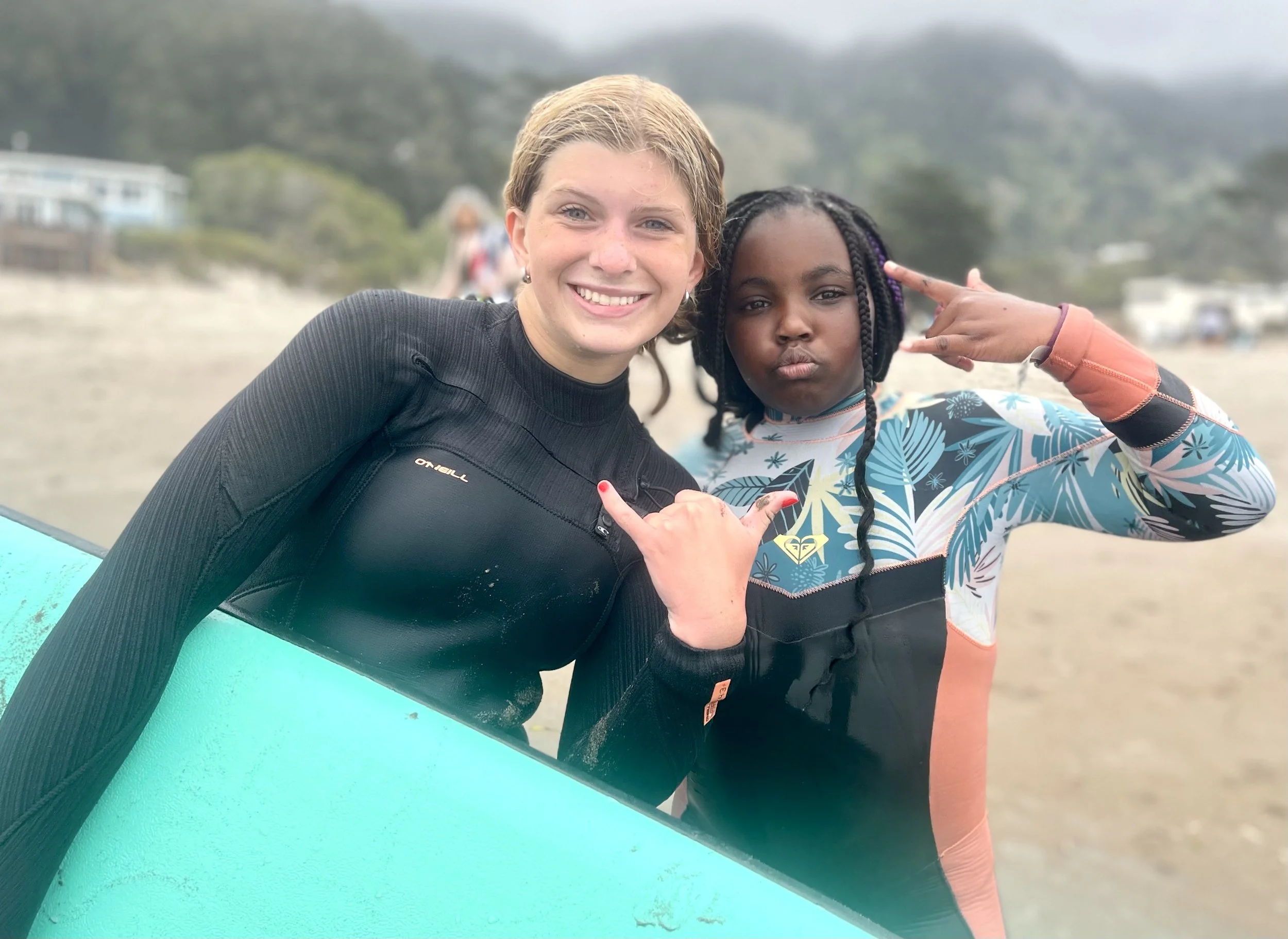 A teenage volunteer and a young participant posing with a surfboard, throwing shaka and peace signs
