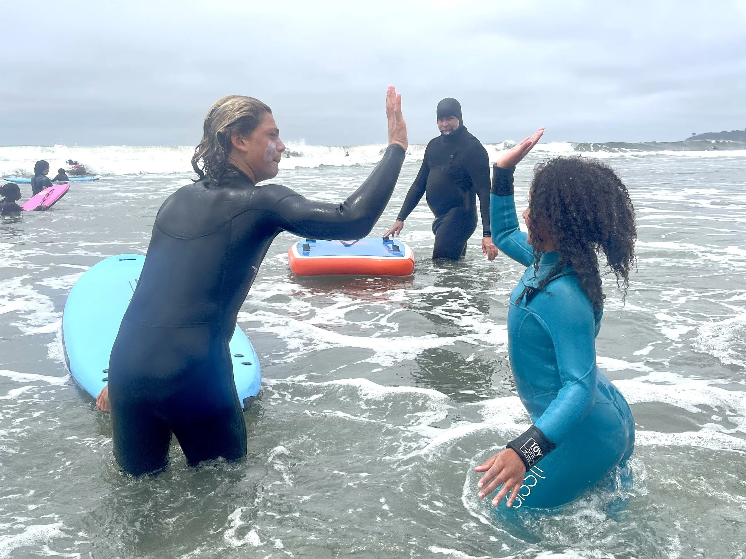 Two people in wetsuits high-fiving in the surf as an instructor looks on