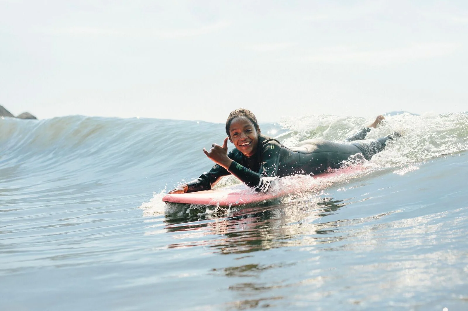 A young girl riding a wave on a pink surfboard, throwing a shaka toward the camera