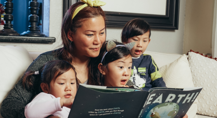 Mother reading a book titled Earth! to her three young children on a couch at home