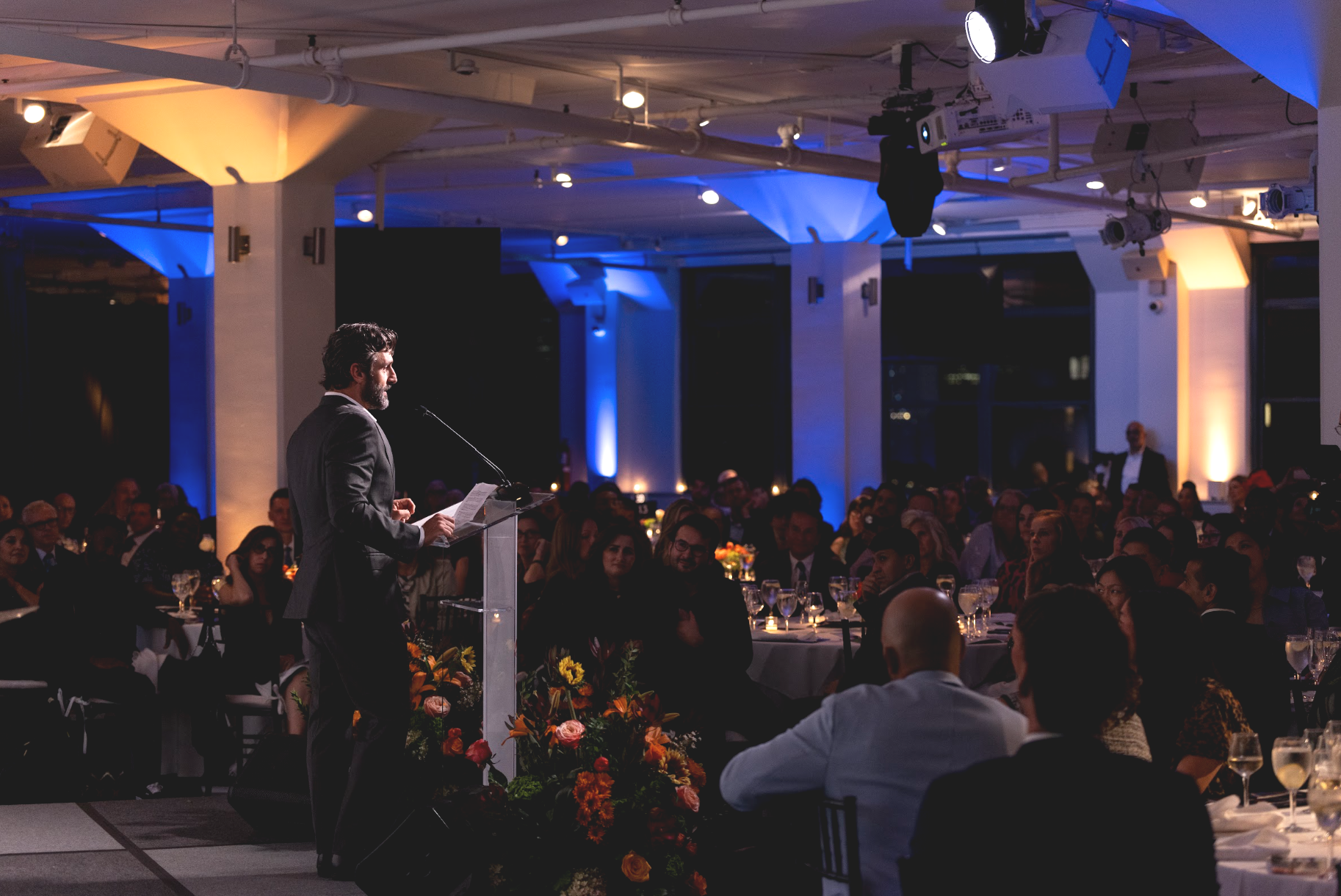 A speaker at a podium addressing a seated gala audience in a blue-lit venue