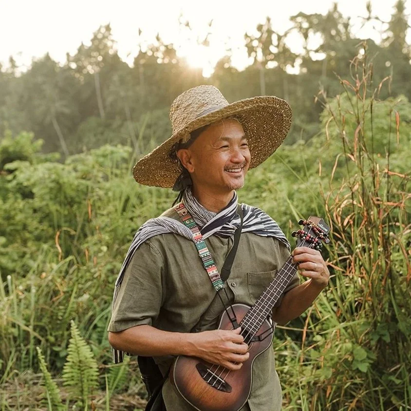 Dr. Home, smiling in a straw hat and holding a ukulele, outdoors in lush greenery at sunset
