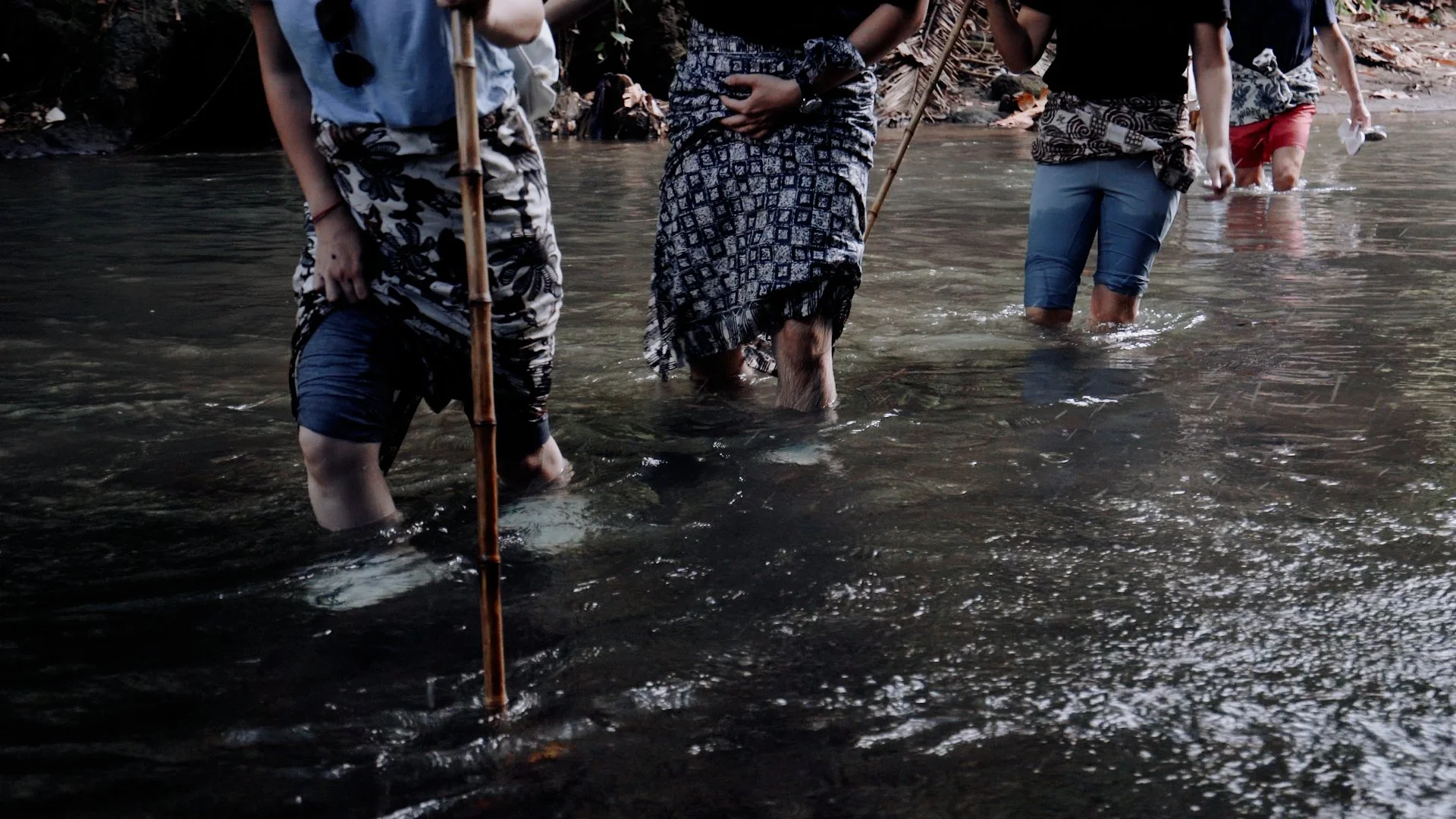 A group of people wading through a shallow Bali stream in sarongs, carrying bamboo poles