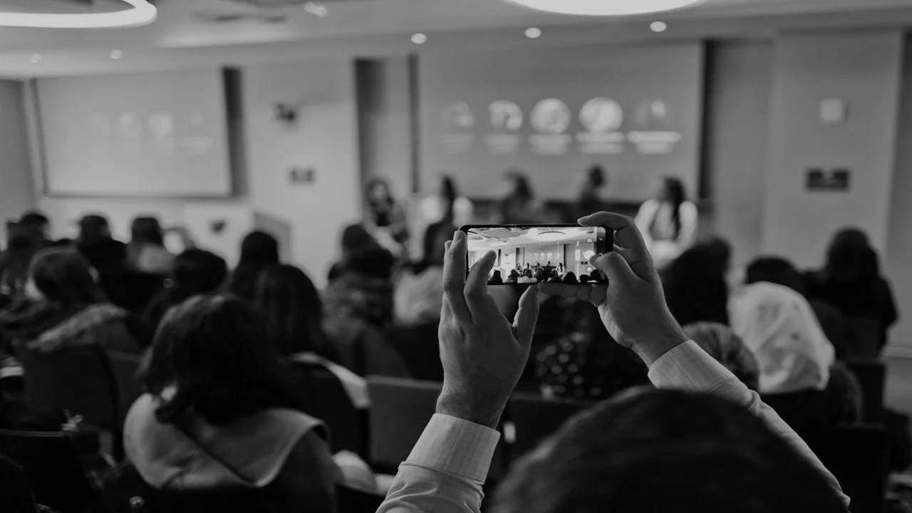 Black-and-white photo of a person photographing a large conference audience with a smartphone