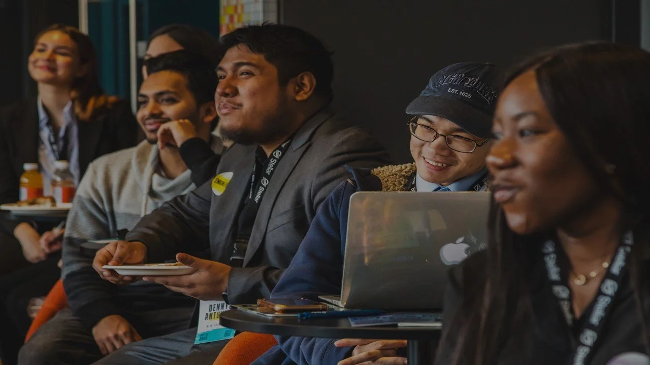Diverse group of young students seated at a Mindot event, smiling and engaged, with lanyards and a laptop