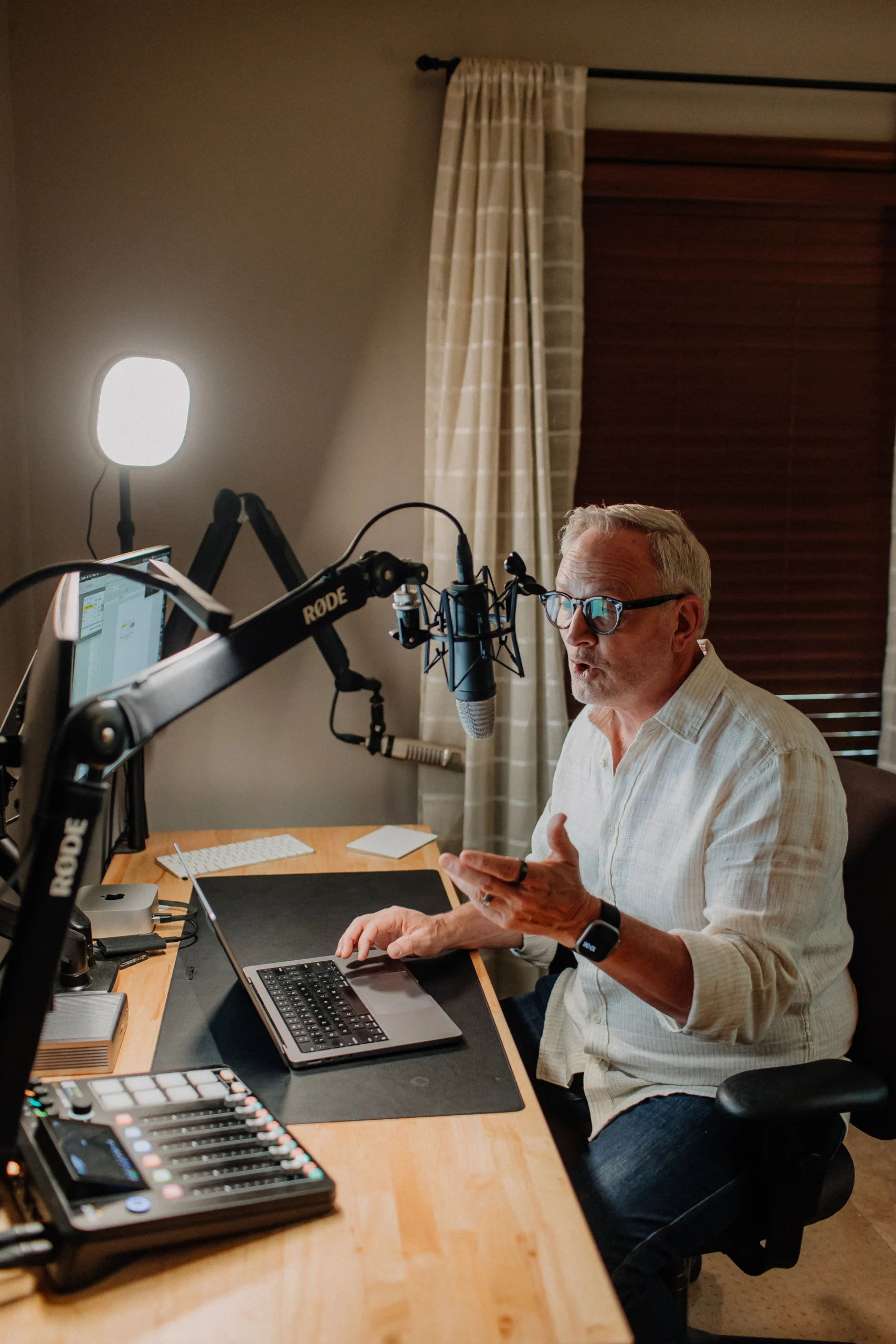A man with glasses speaking at a podcast recording desk with a RØDE microphone