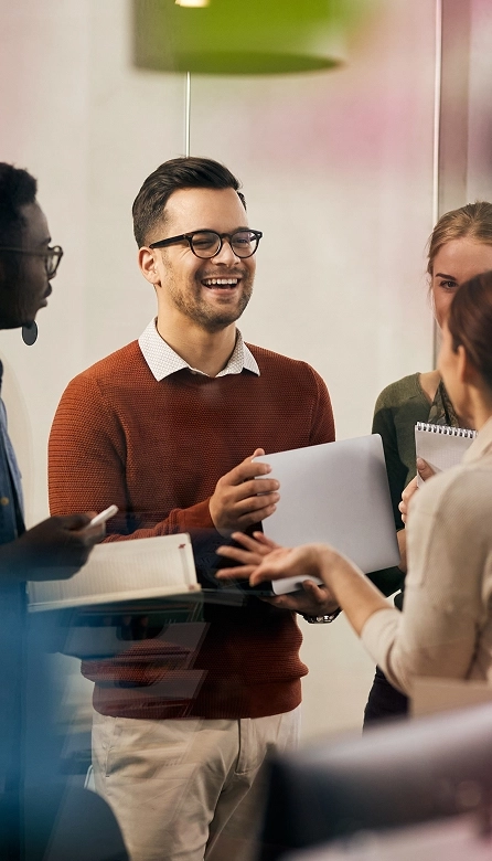 Diverse group of young professionals laughing and collaborating in an office, one man in a red sweater holding a laptop