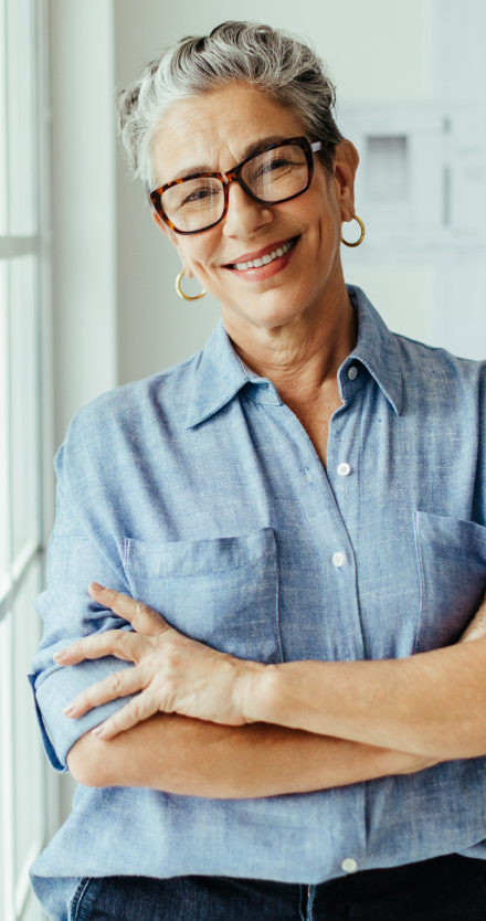 Smiling professional woman with grey hair, tortoiseshell glasses and a chambray shirt, arms crossed