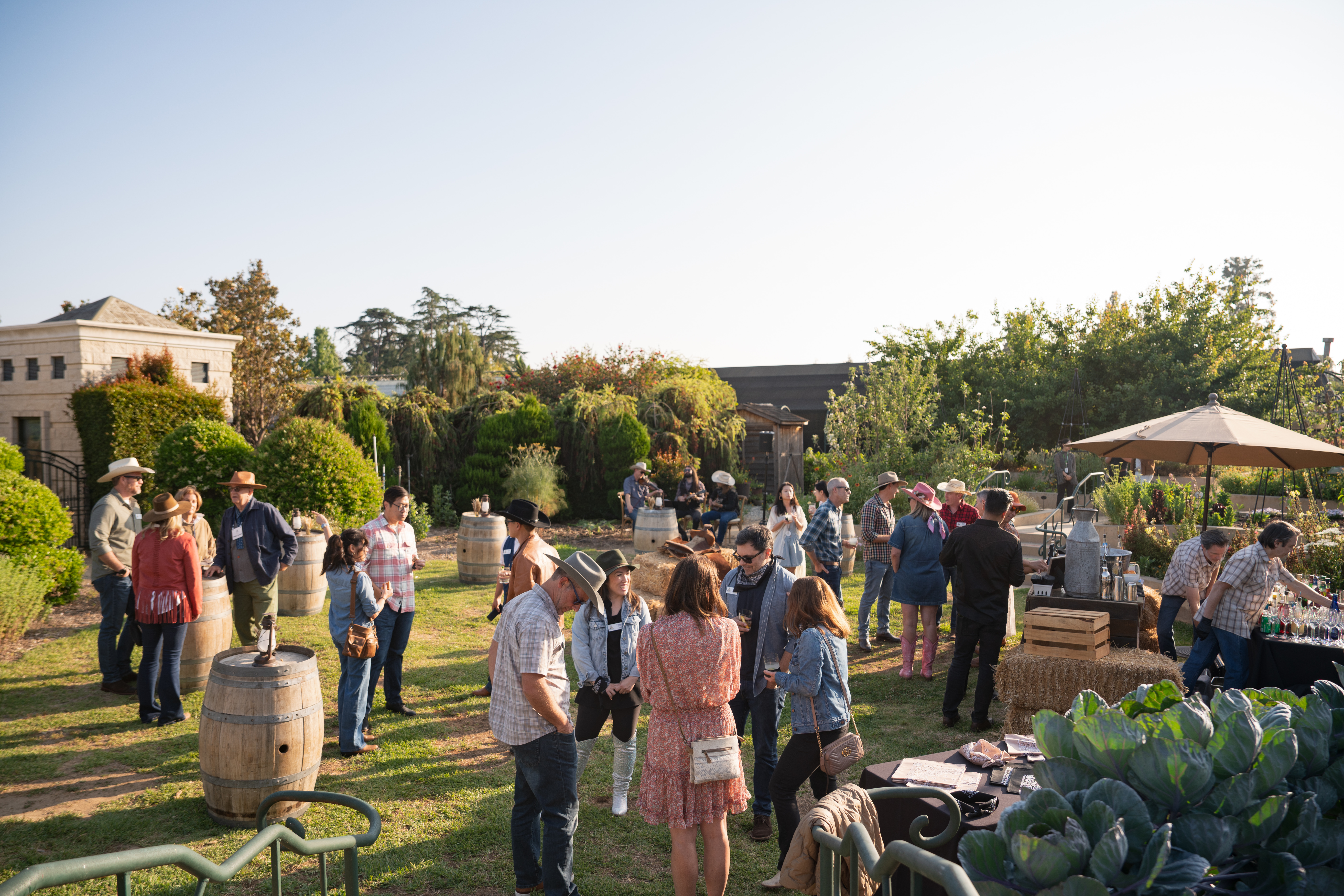 Wide-angle outdoor western-themed corporate event with guests mingling among wine barrels and hay bales at golden hour