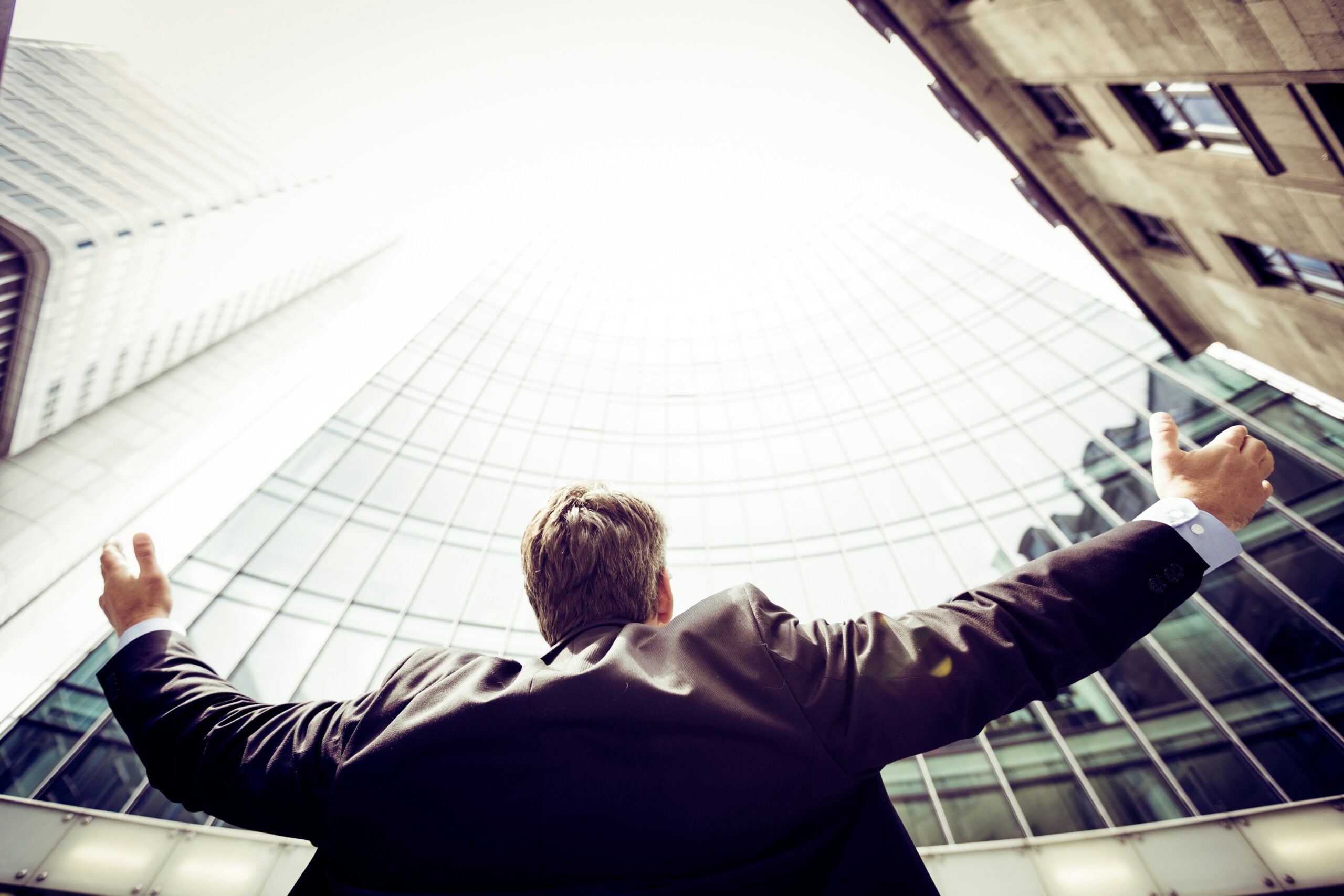 Businessman in a suit facing glass skyscrapers with arms raised in triumph