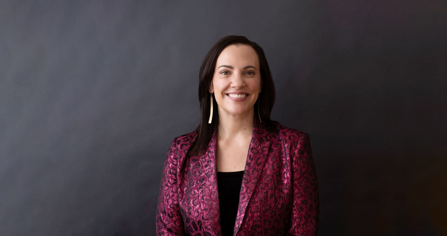 Natalie Micale smiling in a magenta patterned blazer against a dark grey background
