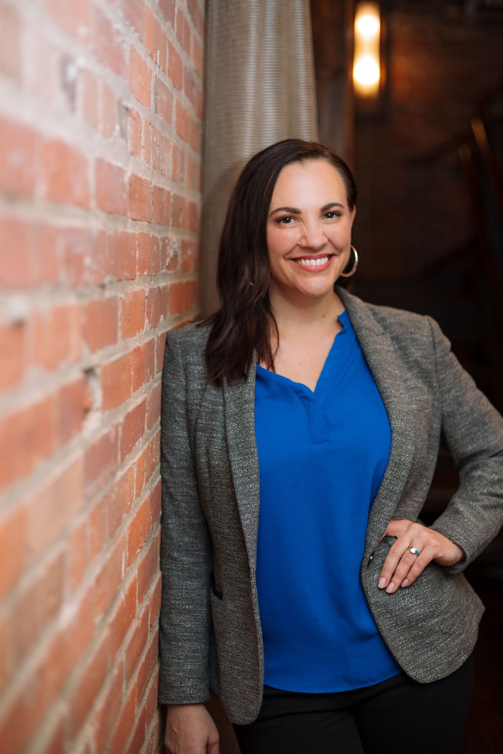 Natalie Micale in a blue blouse and grey blazer leaning against a brick wall