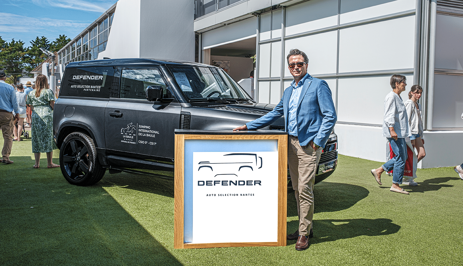 Man in a blue blazer standing beside a Defender-branded wooden trade-show counter at an outdoor event