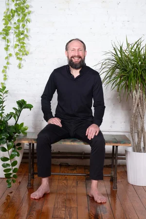 Conor Yates — bearded instructor in black seated on a bench against white brick with plants