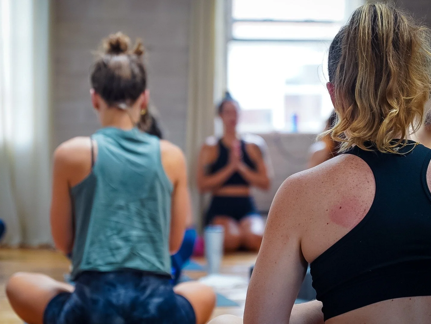 Students from behind facing a yoga instructor in prayer pose at the front of a studio class