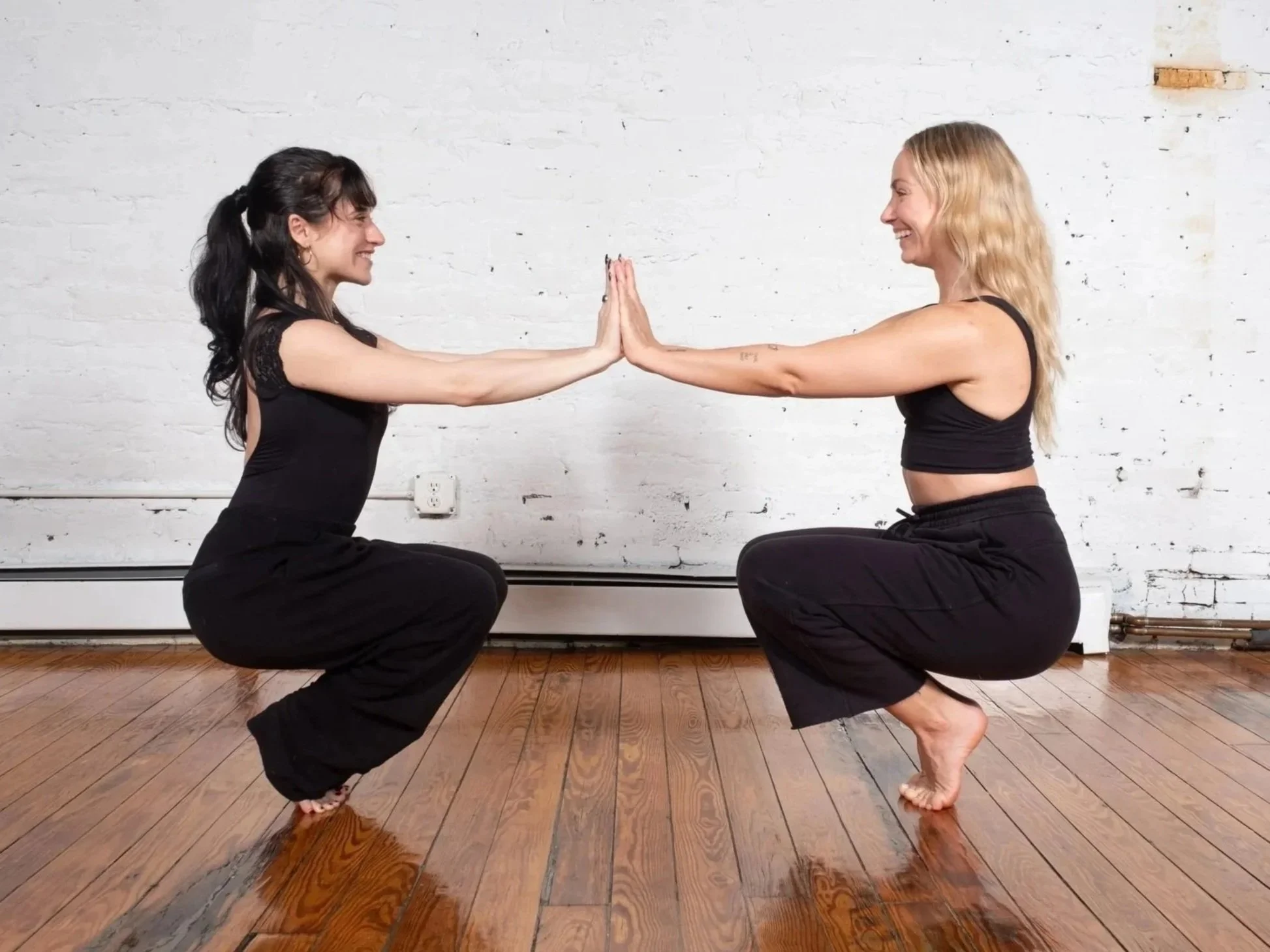 Two women facing each other in a partner eagle-squat pose, palms pressed together, in the white brick studio