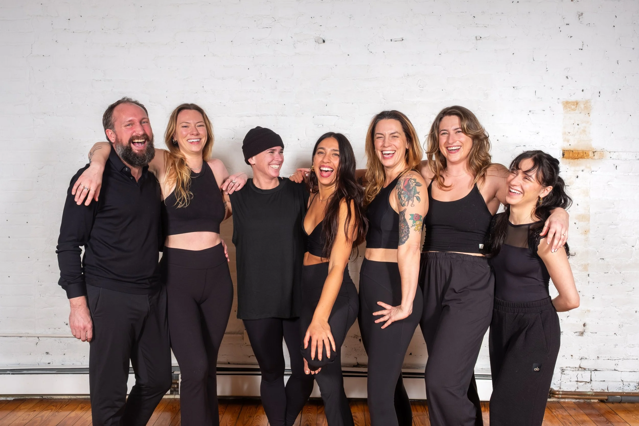Seven yoga instructors laughing together arm-in-arm against a white brick wall