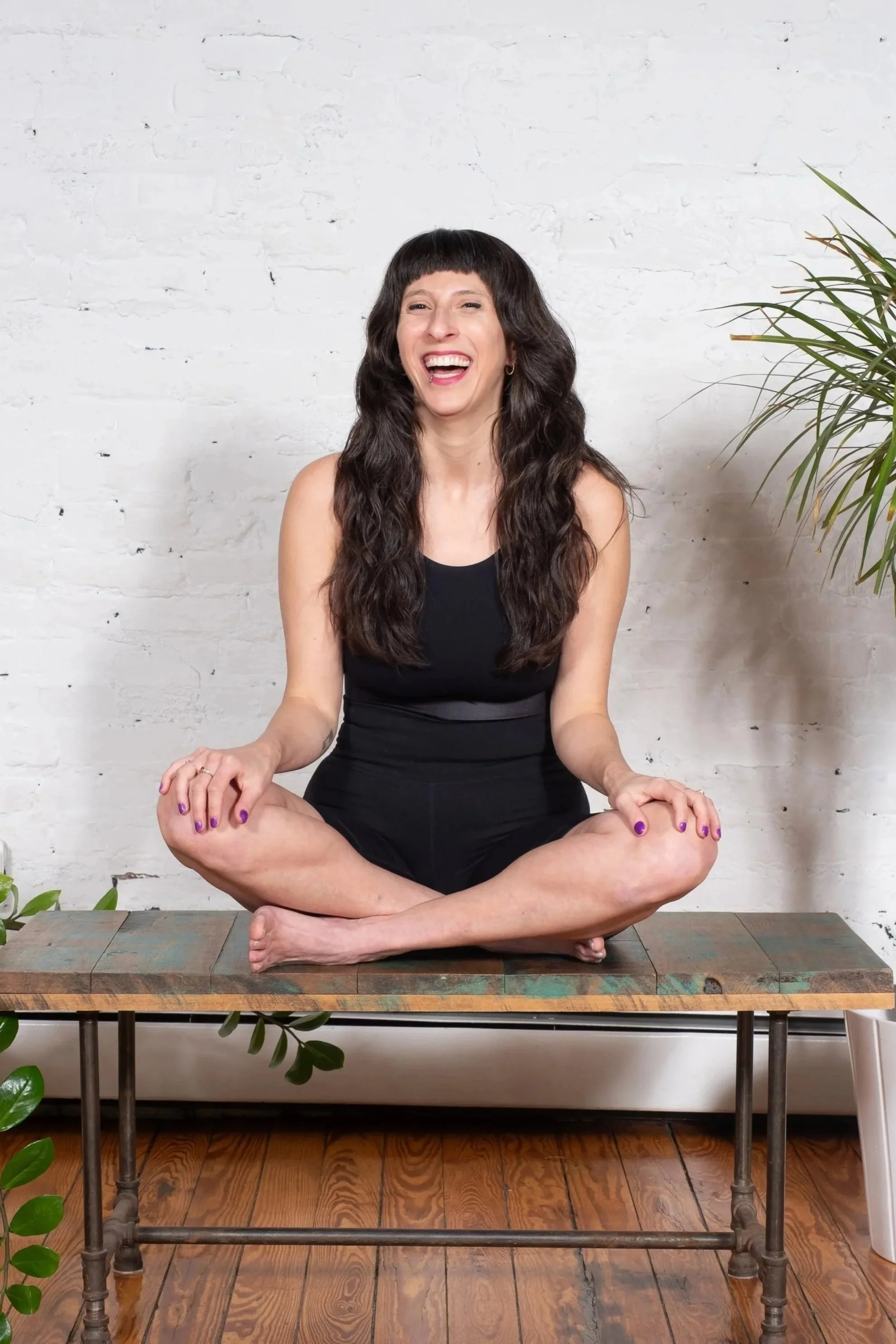 Liana Rosenberg — instructor with bangs in a black bodysuit seated cross-legged on a bench, laughing