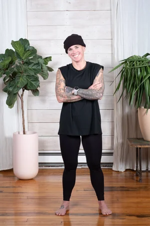 Be Leigh — instructor in a black beanie and sleeveless shirt, arms crossed, against a wood-panel backdrop