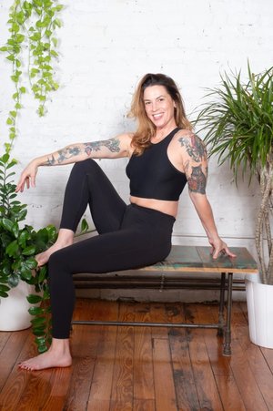 Keela Williams — tattooed instructor in black seated on a bench against white brick with plants