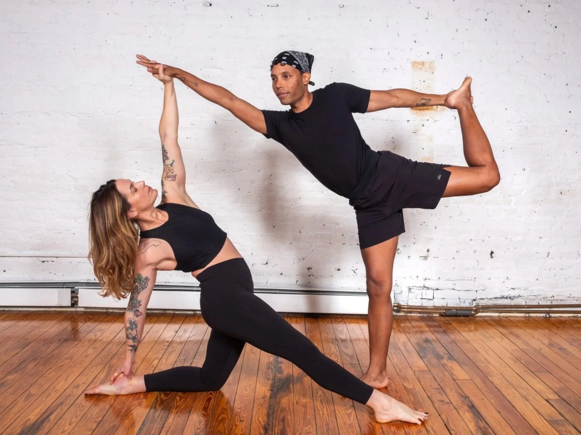 A man in dancer's pose and a woman in a low lunge reaching up, practicing partner yoga in the white brick studio