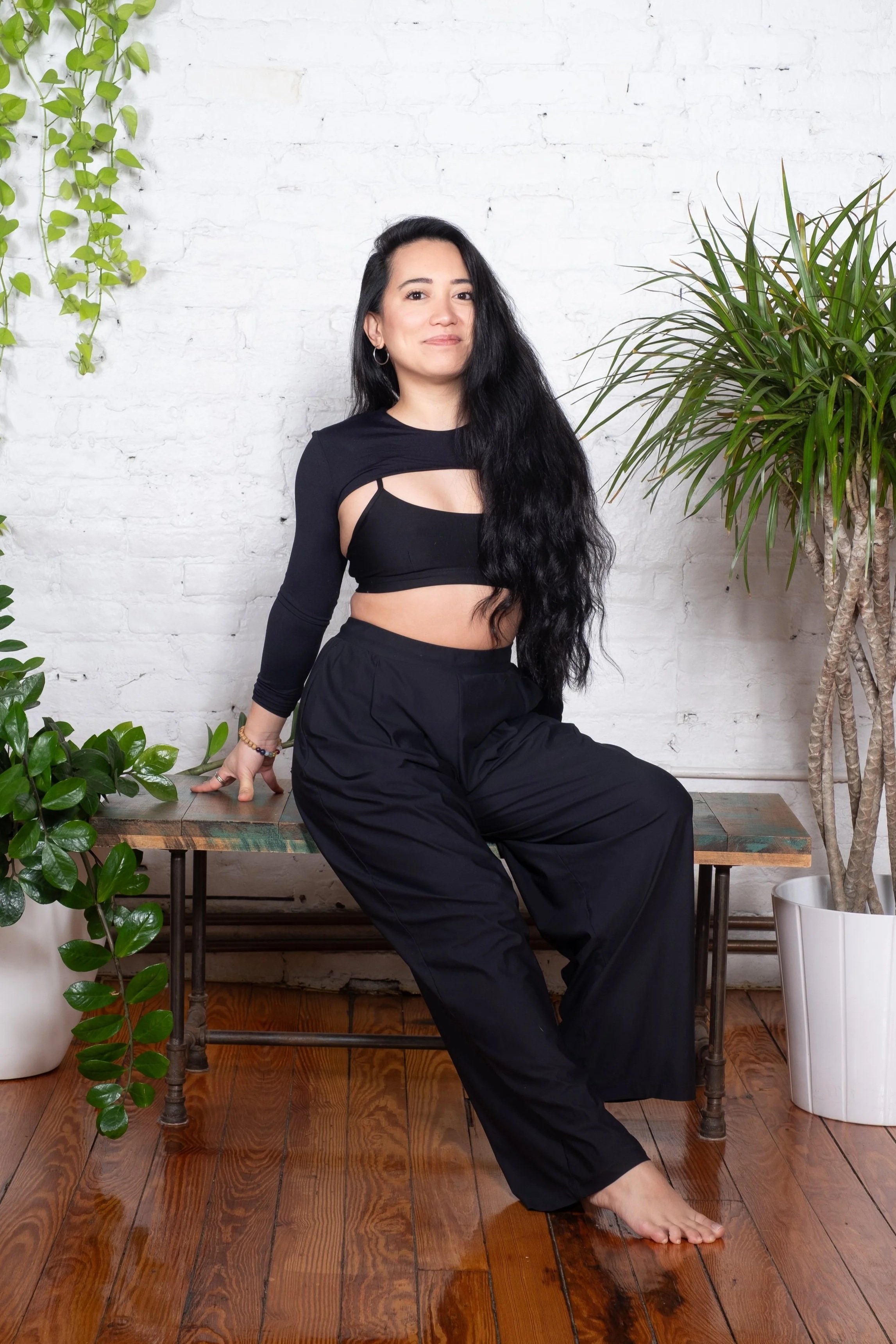 Samantha Casuga — instructor with long dark hair in a black cut-out top seated on a bench with plants