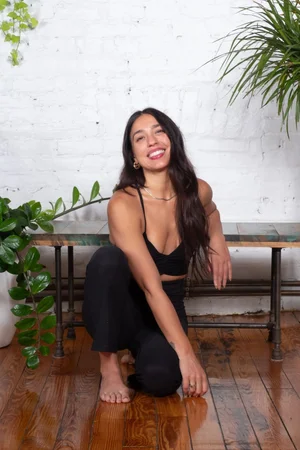 Lulu Soni — instructor with gold hoop earrings in black, seated on the floor against white brick, smiling