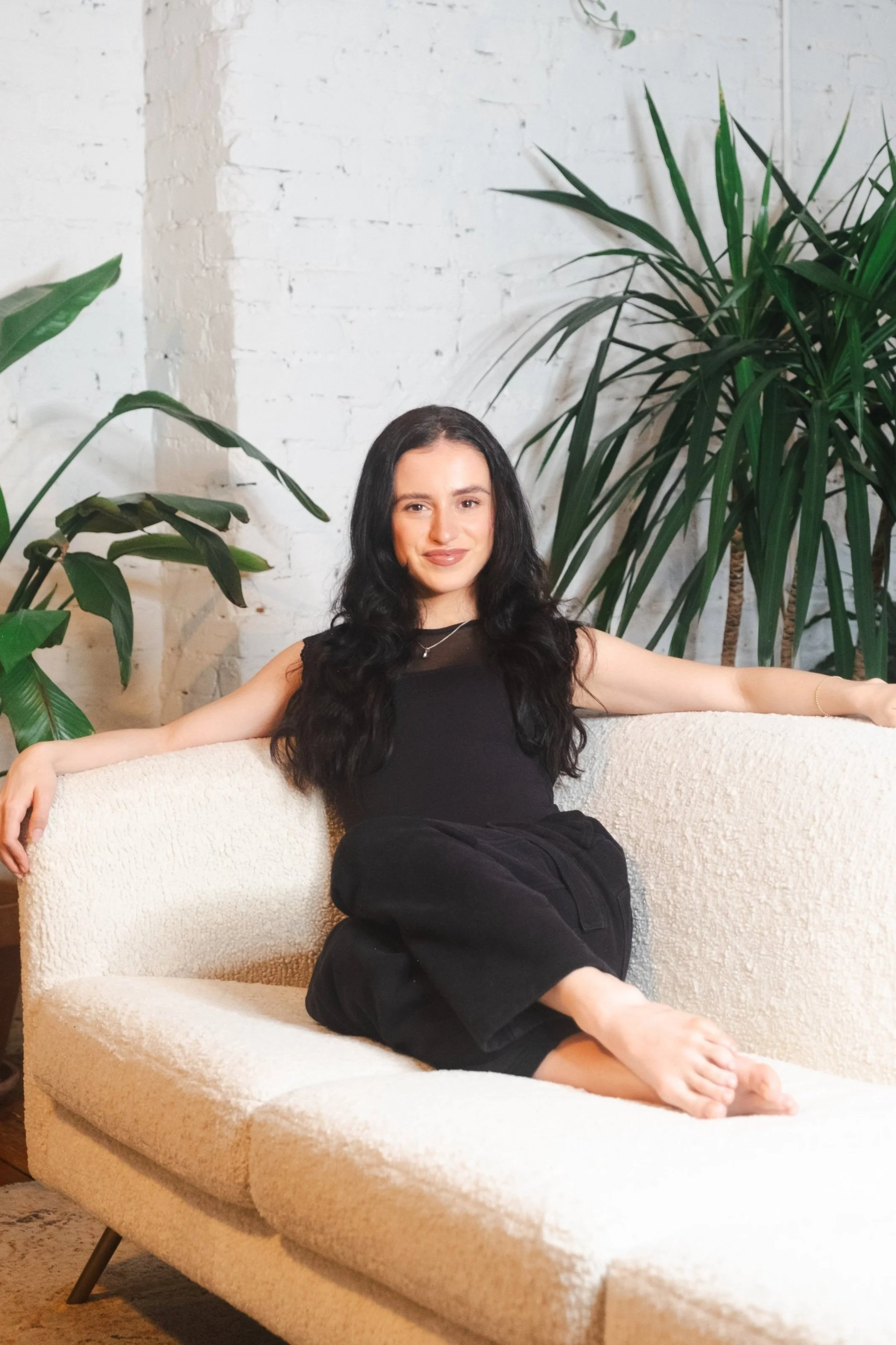 Julia Padavona — instructor in a black sleeveless dress seated on a cream boucle sofa surrounded by plants