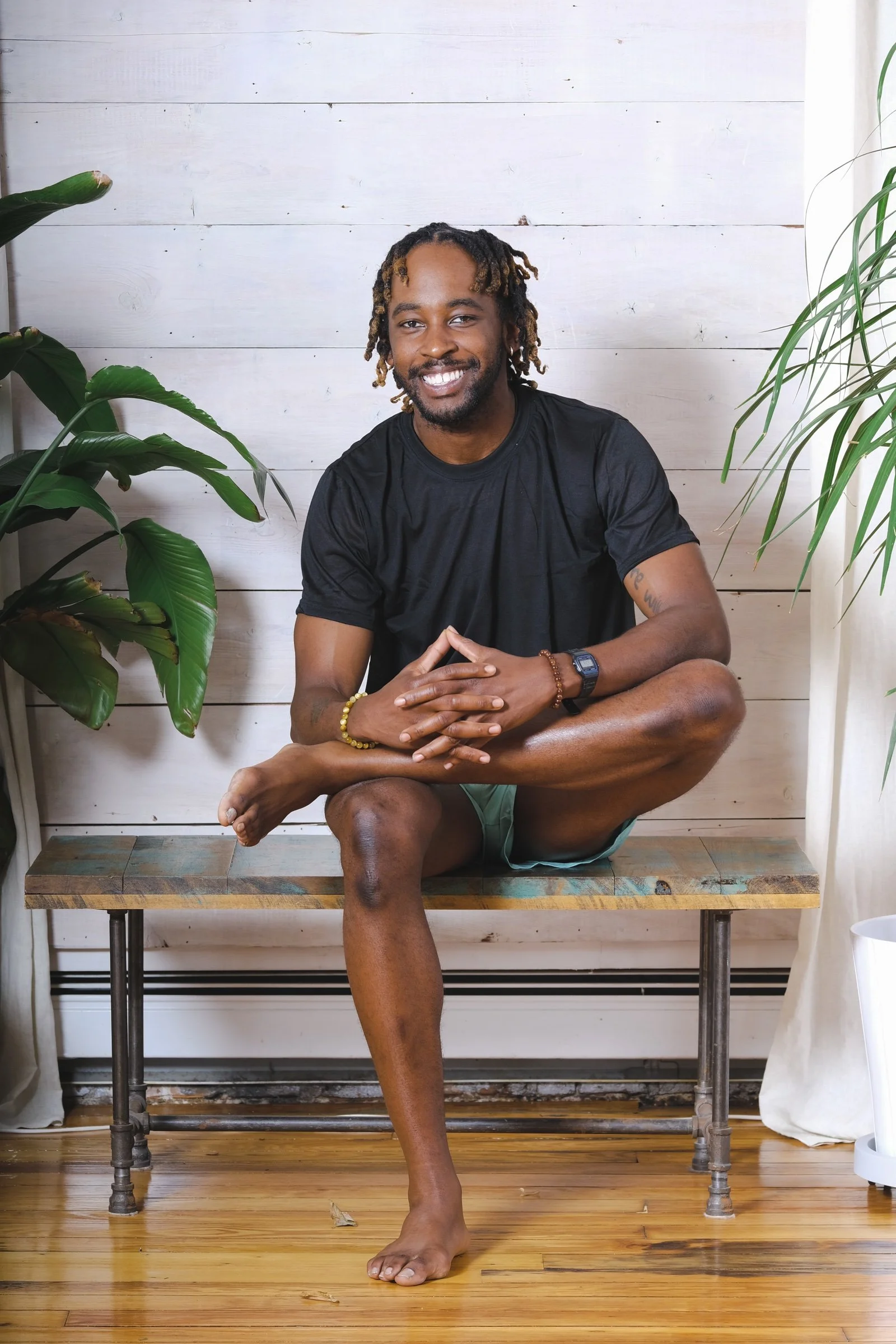 Quazzy Herd — instructor with locs in a black t-shirt seated cross-legged on a bench, smiling