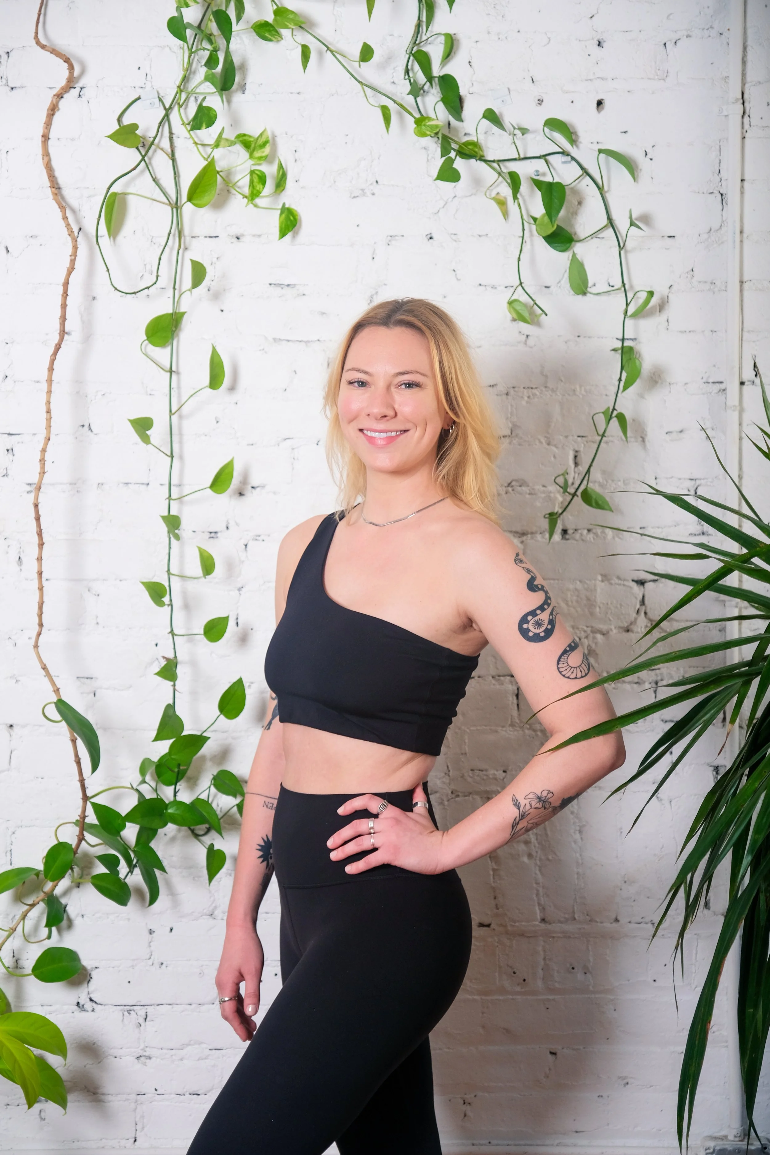 Emma Henry — blonde instructor in a black one-shoulder crop top against a white brick wall with vines