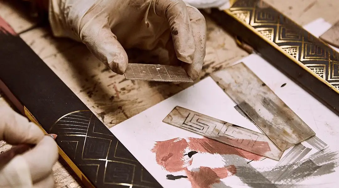 Gloved hands applying gold leaf to an ornate picture frame on a worktable