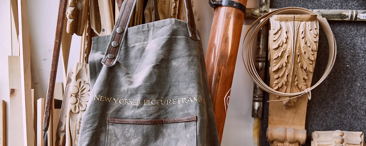 New Yorker Picture Frames branded canvas apron hanging in the shop among wood mouldings and carved ornaments