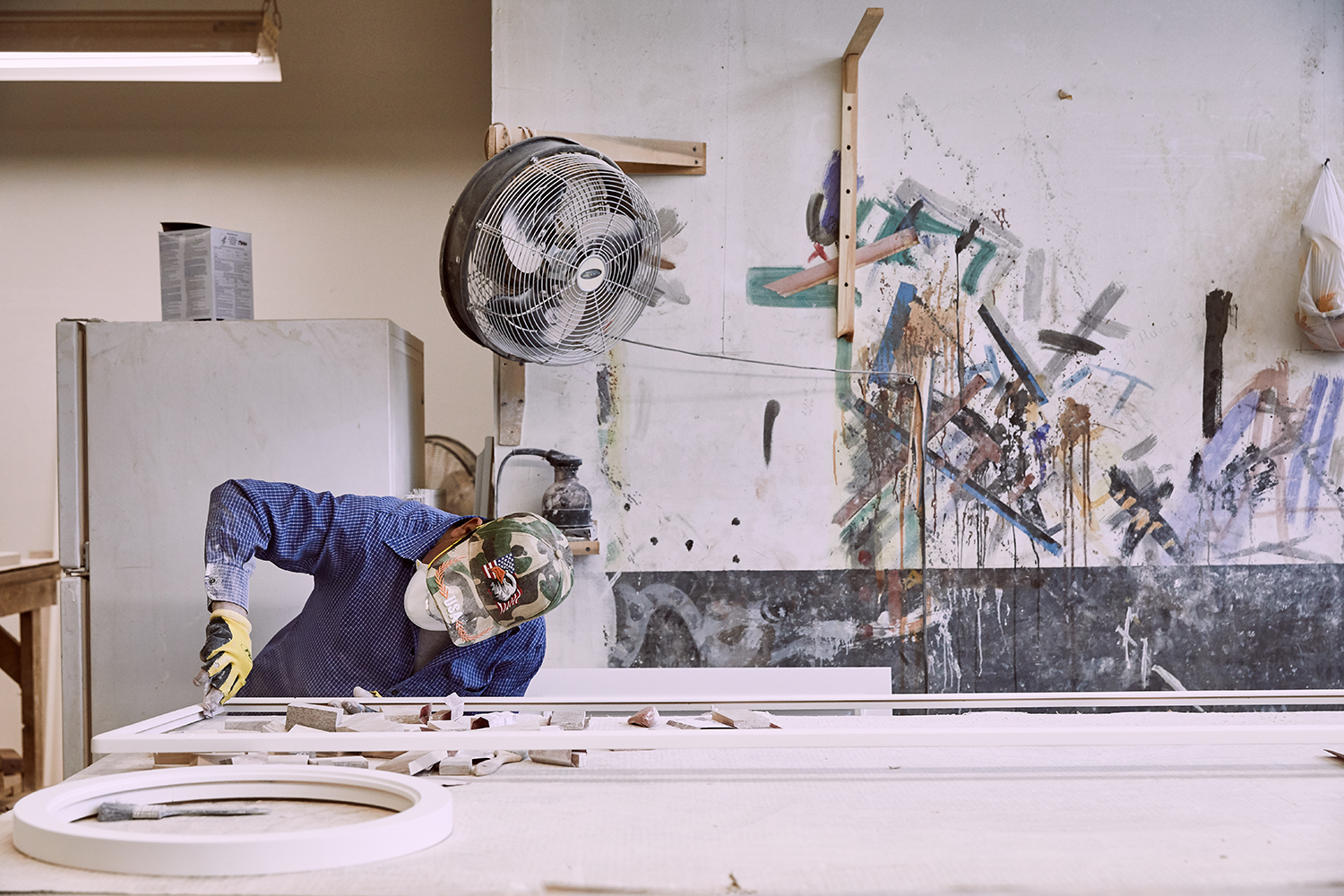 Craftsman in cap and gloves working picture frame mouldings, abstract painting on the wall behind
