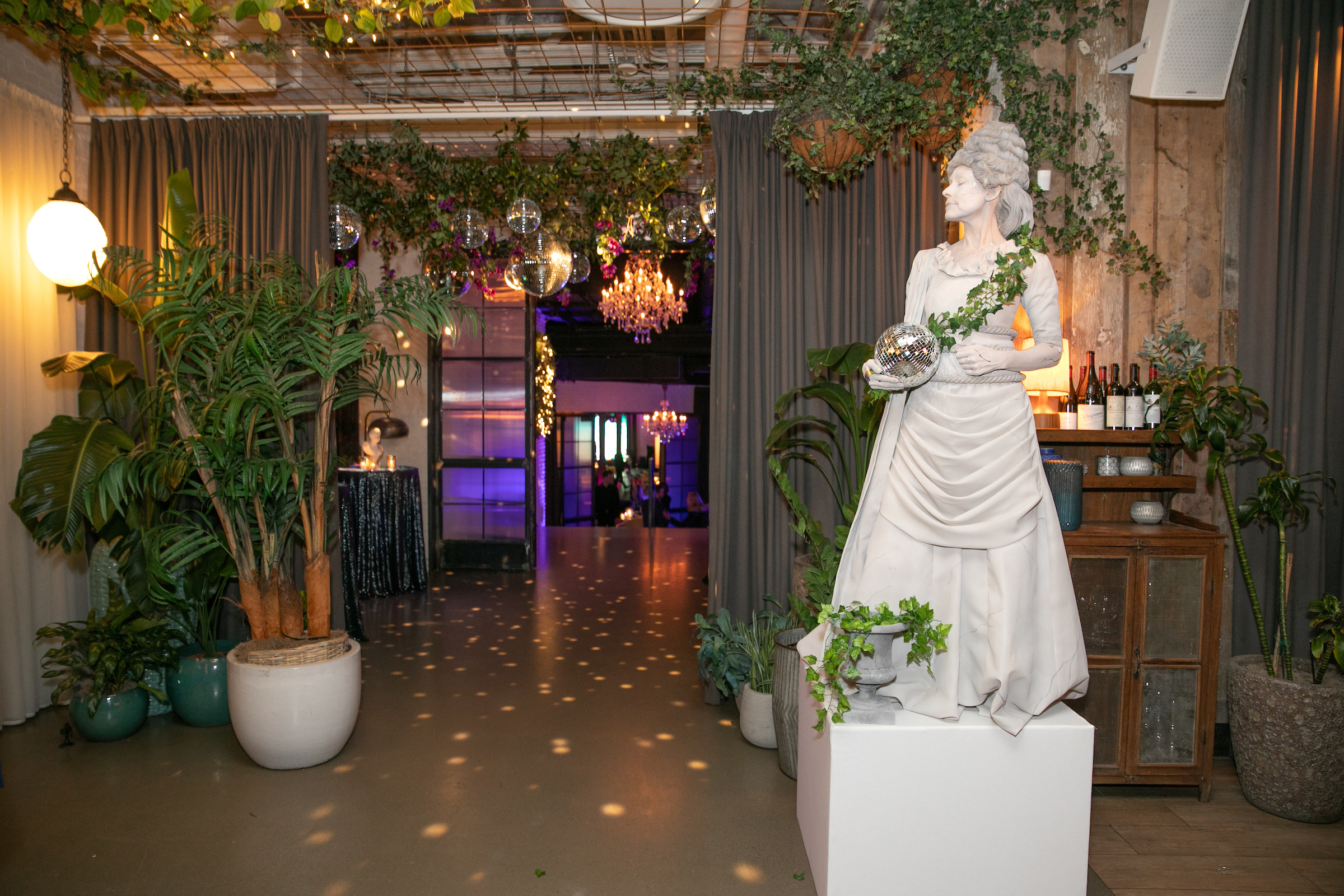Gala venue entrance with a white classical statue holding a disco ball, lush greenery, chandeliers and draped curtains