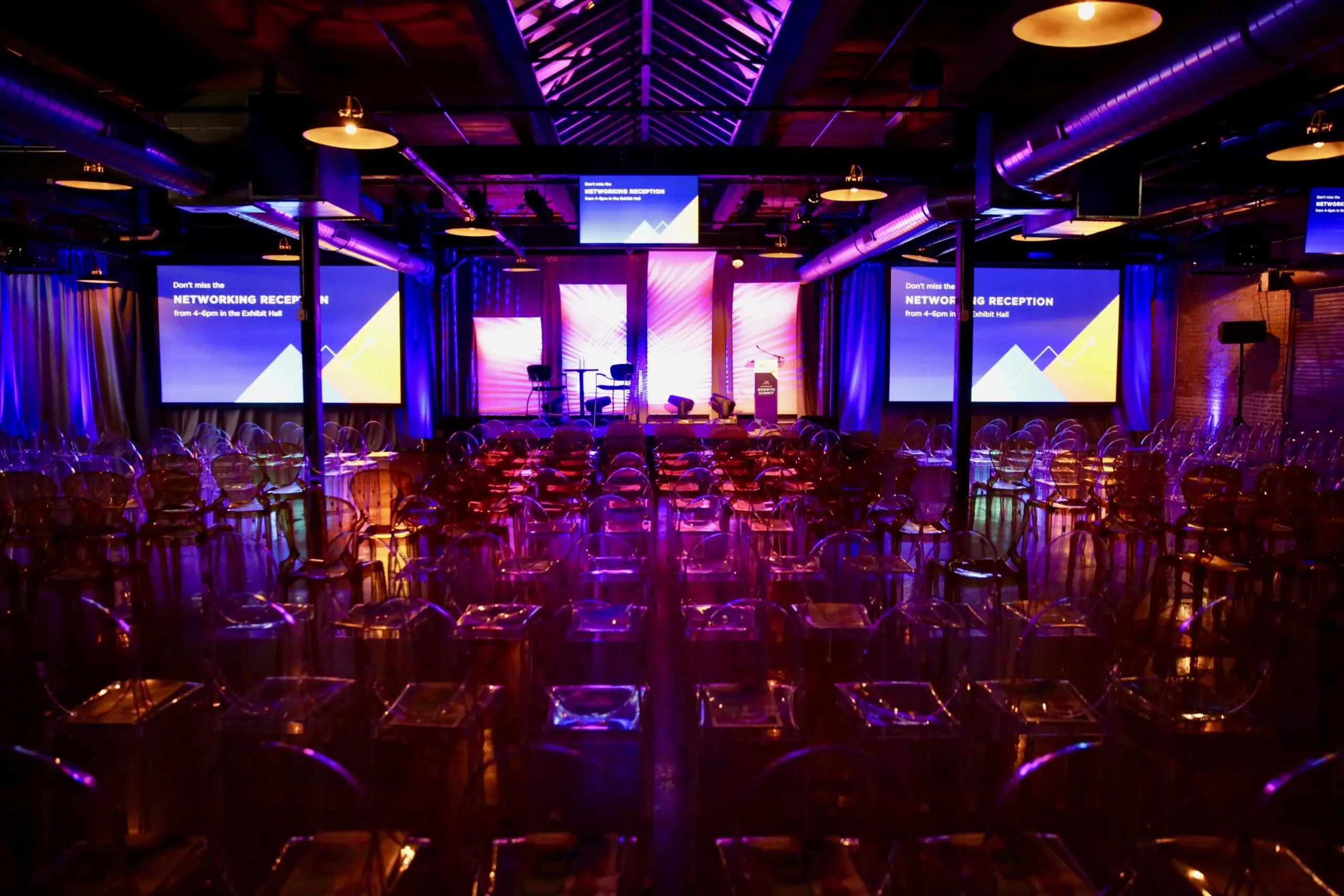Conference space with purple uplighting, ghost chairs, multiple LED screens and a center stage