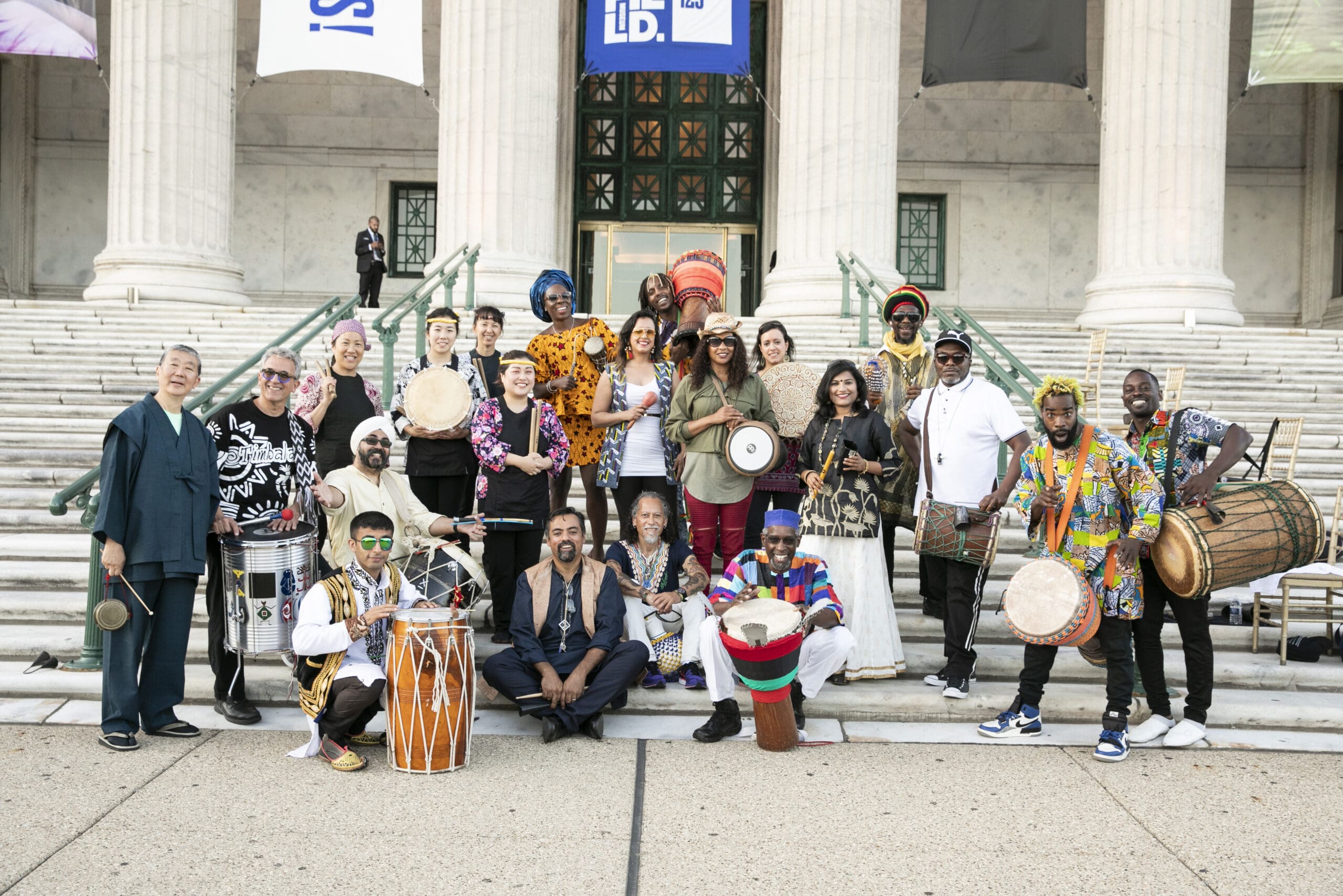 Group of multicultural drum performers posing on the steps of a neoclassical building at an outdoor event