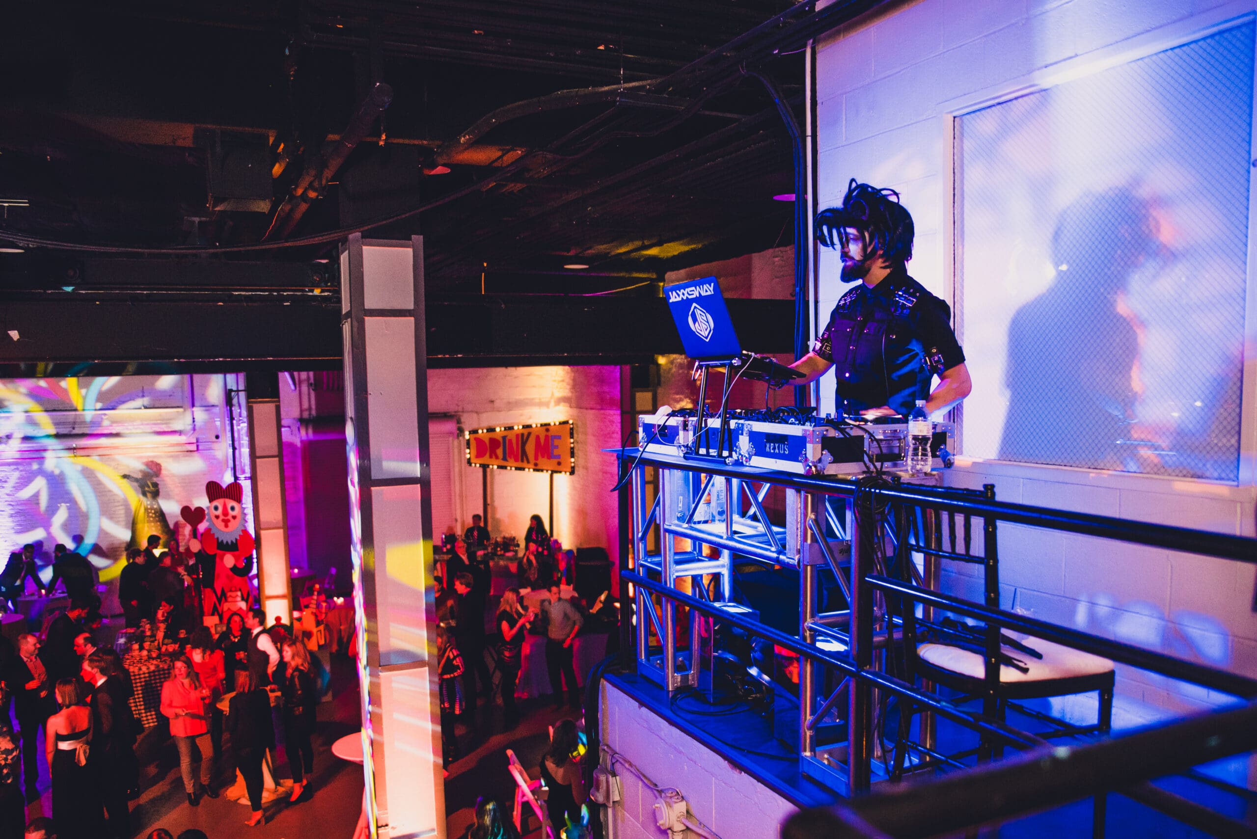 DJ and vocalist performing on an elevated platform at a colorful indoor corporate event with a crowd below and neon signage