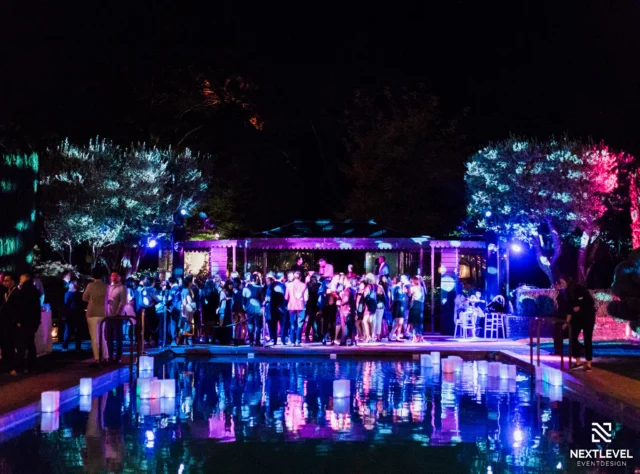 Outdoor nighttime poolside event with guests under blue and purple uplighting