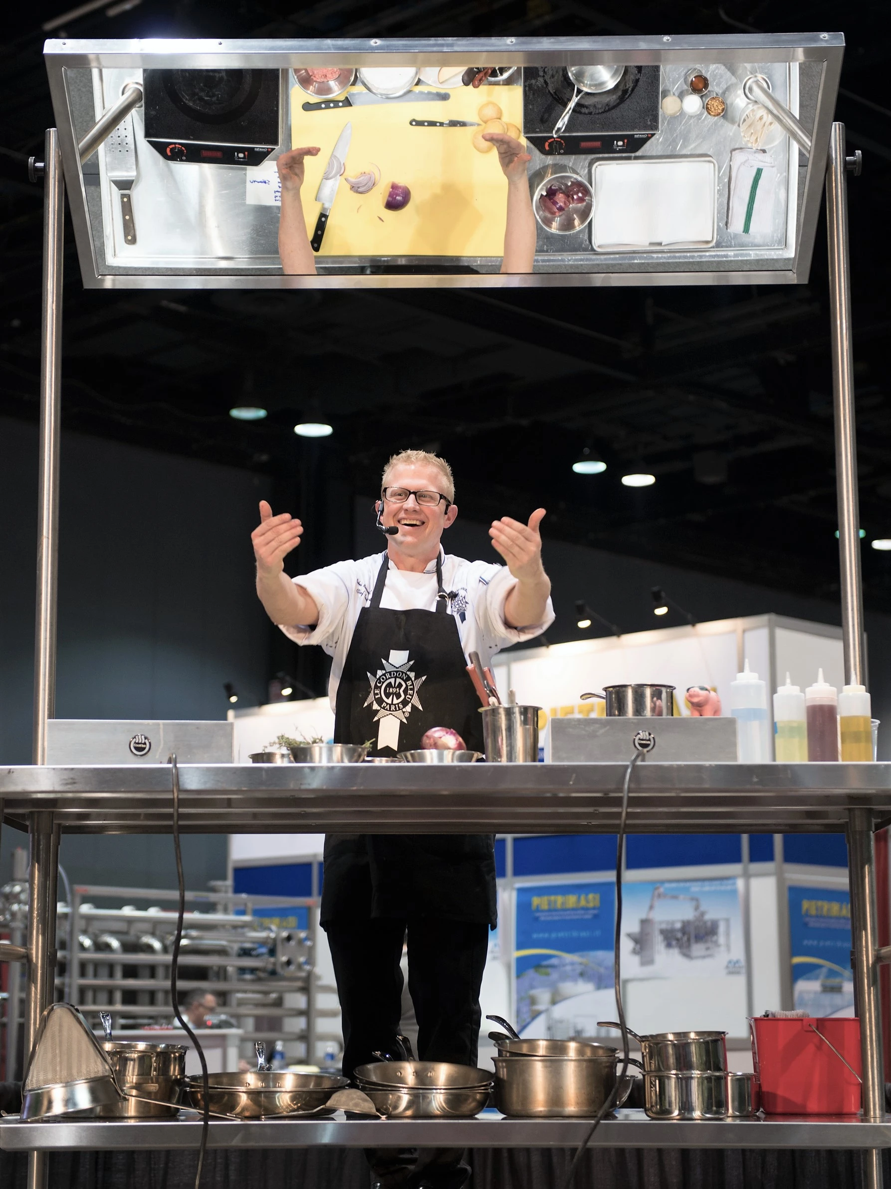 Chef in a Le Cordon Bleu apron running a product launch cooking demonstration at an expo with an overhead mirror display