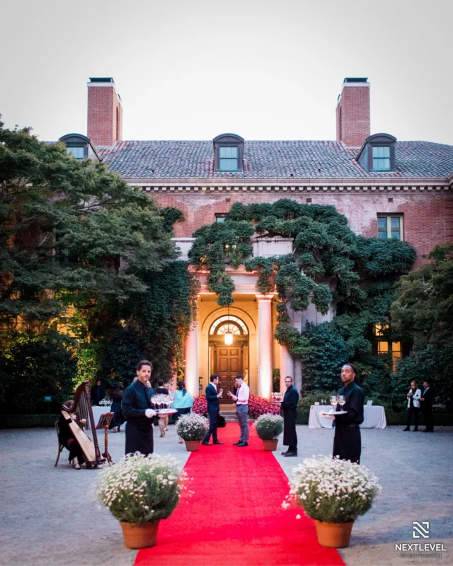 Elegant estate exterior with a red carpet entrance, servers with champagne trays and a harpist