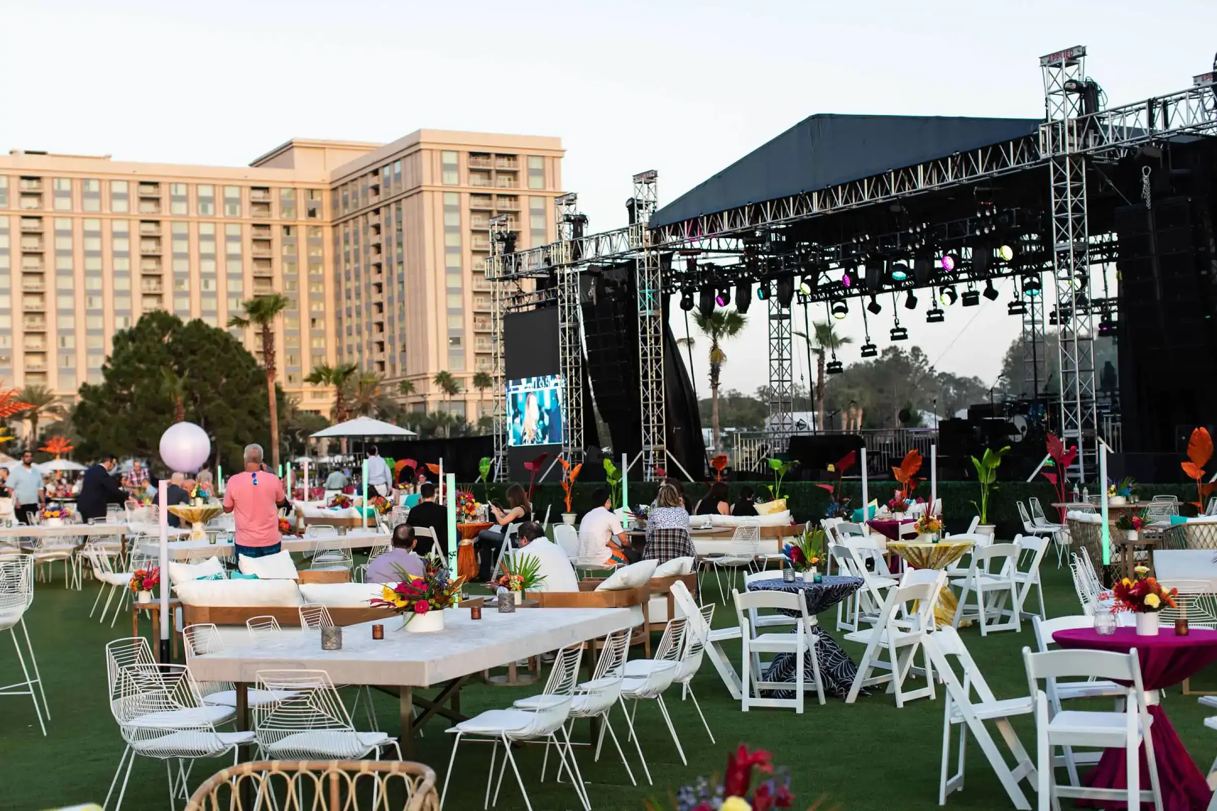Outdoor incentive concert on a grass field with a large stage, colorful lounge seating and palm trees at dusk