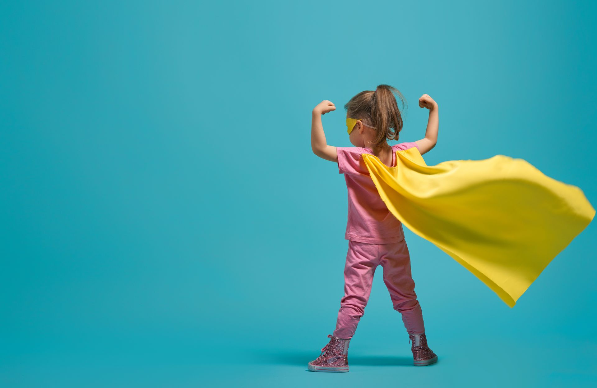 A young girl in a pink shirt and flowing yellow superhero cape and mask, arms raised and flexing.