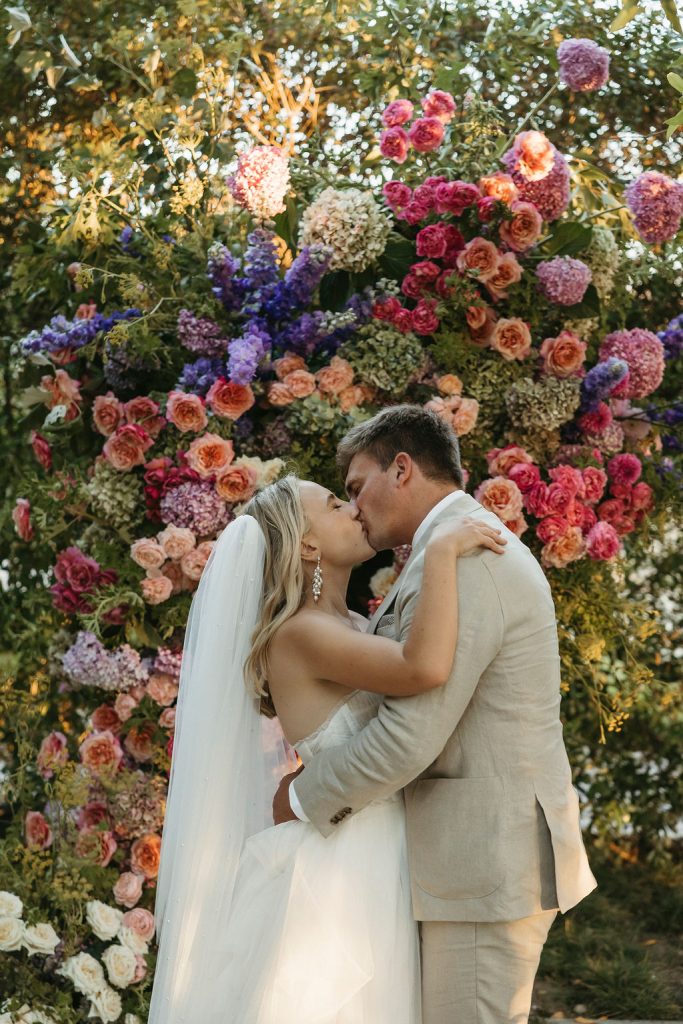 Anja & AJ kissing under a colourful floral arch at Belair