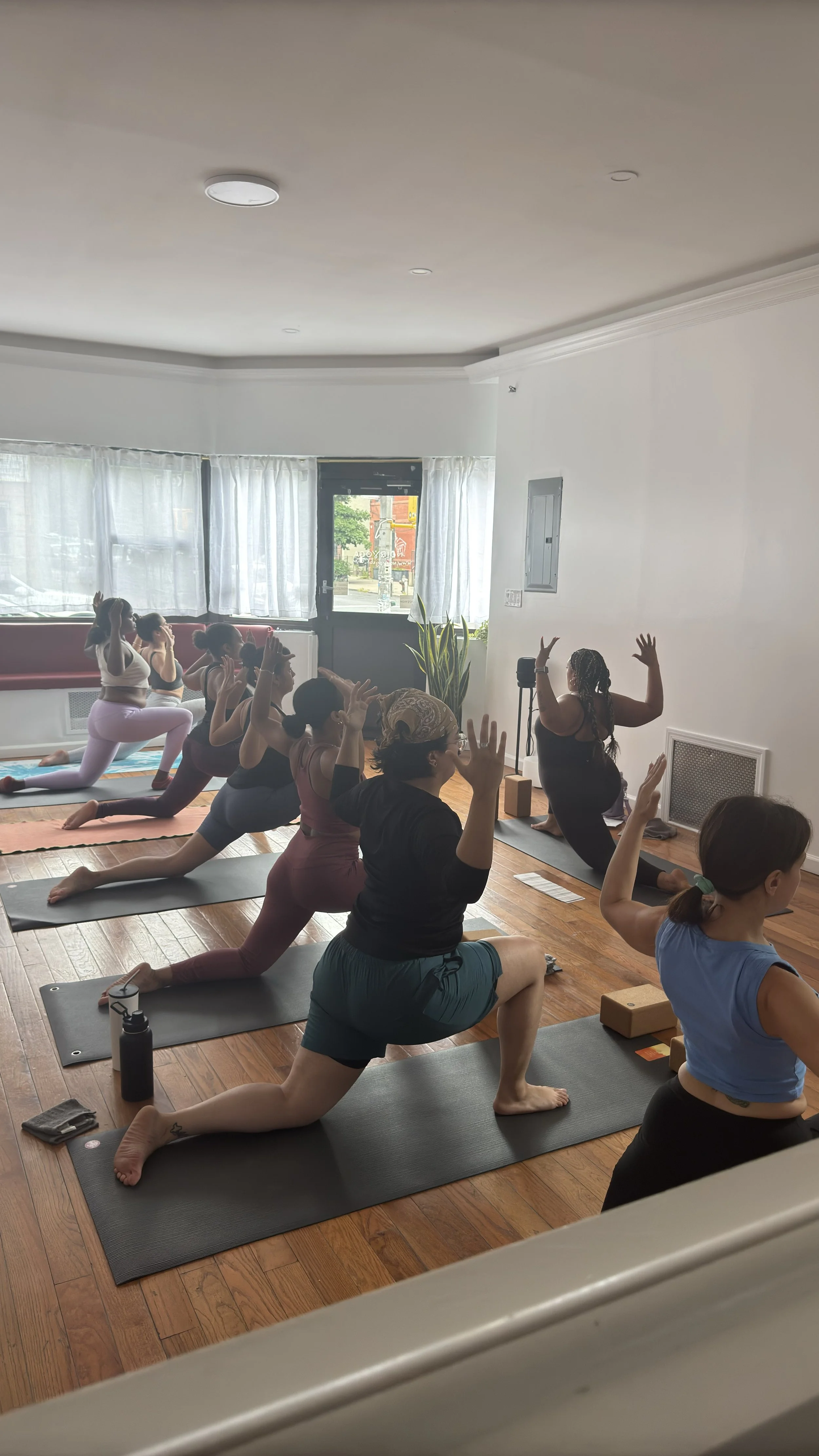 Class is in session at Noir Yoga — students on mats in low lunge, bright street-facing studio