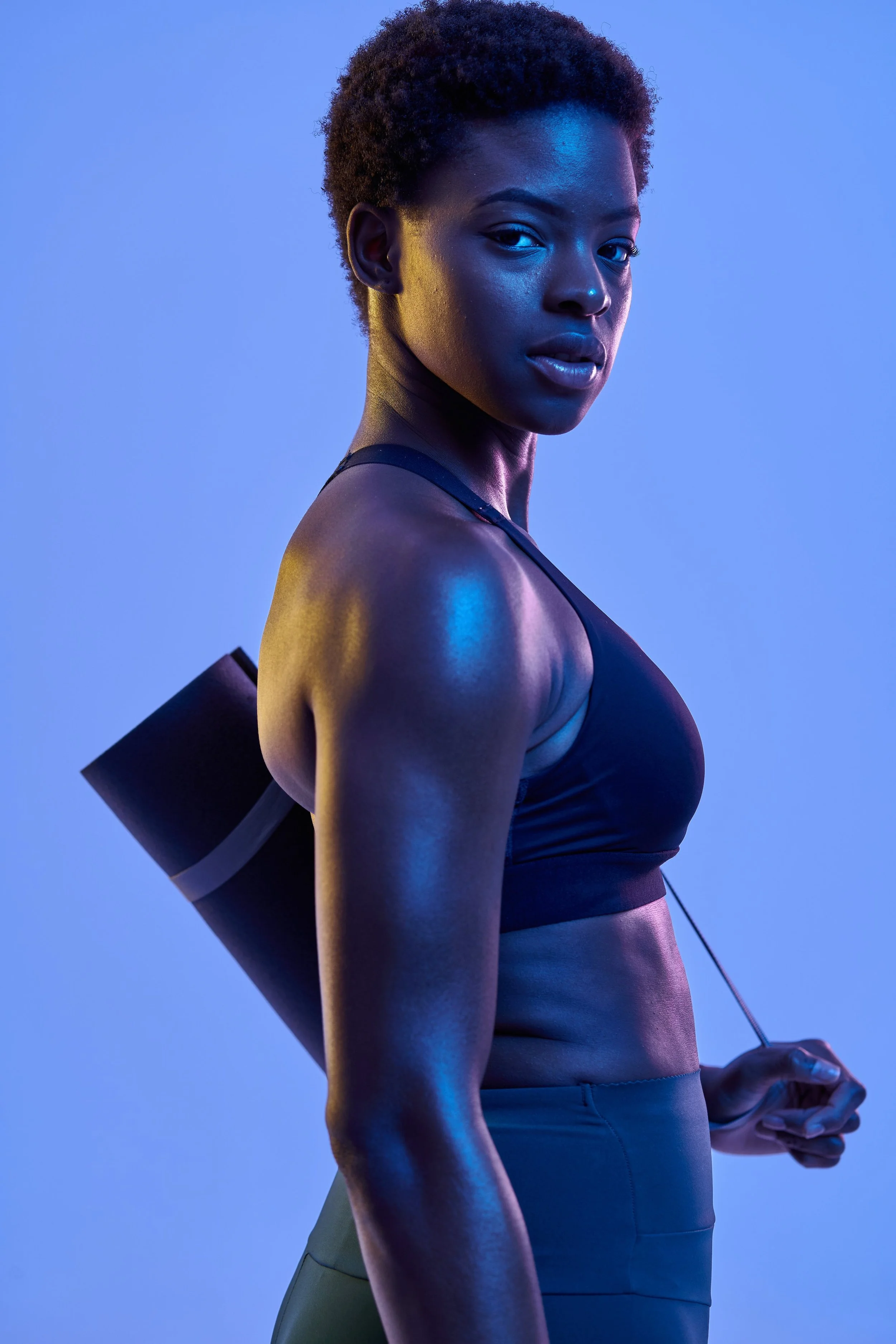 A Black woman in a sports bra holding a rolled yoga mat under dramatic blue studio lighting