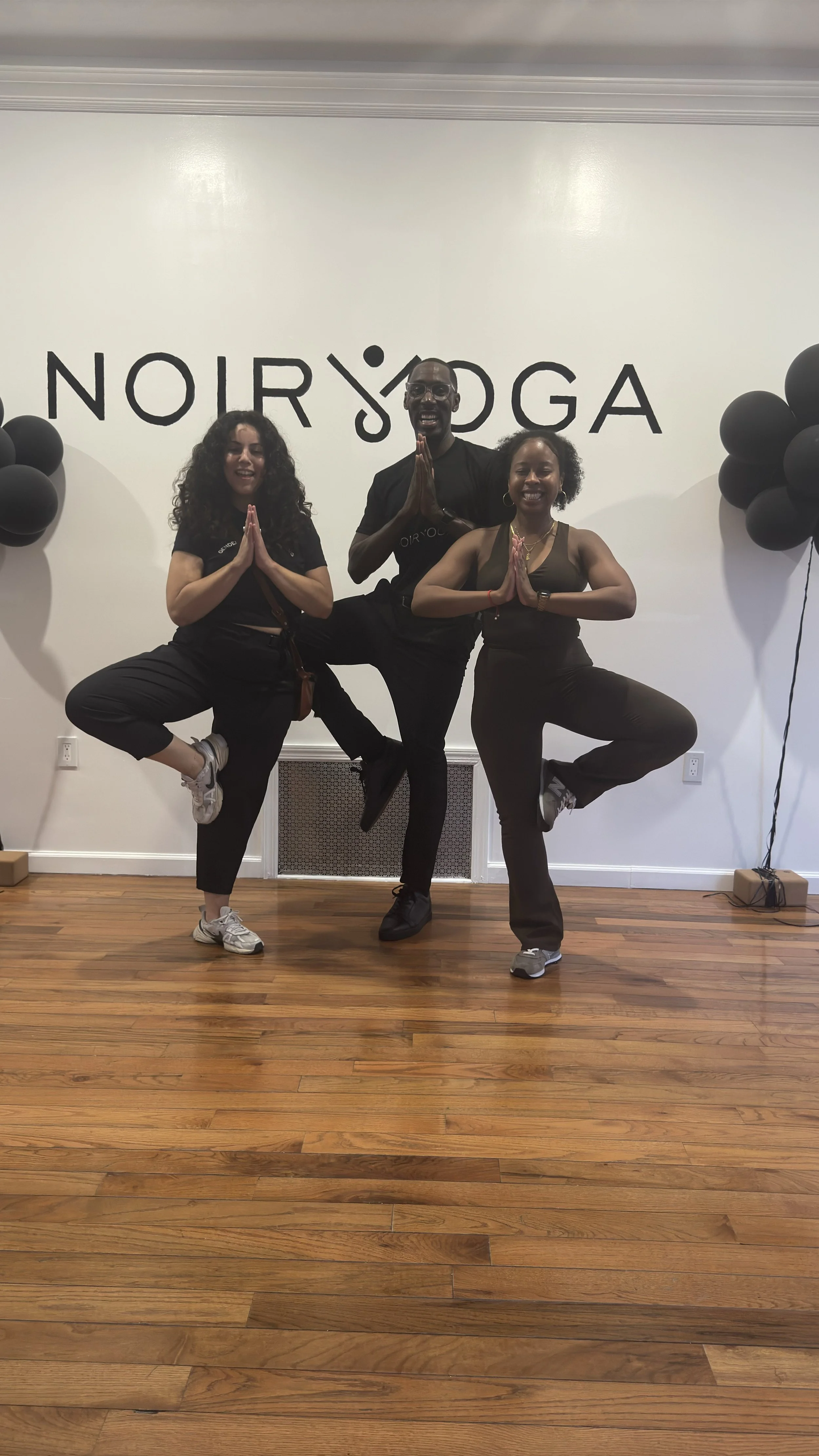Three yoga instructors doing tree pose in front of the black Noir Yoga logo on a white wall